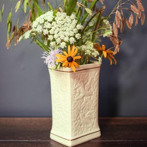 May include: A cream-coloured ceramic vase with a square base and embossed bird and foliage patterns, featuring a thin gold rim. The vase is filled with a bouquet of white Queen Anne's lace, orange coneflowers, and tall, slender brown grasses.