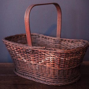 May include: A brown woven wicker basket with a curved handle. The basket has a rounded shape and is made of interwoven strips of wood. The handle is also made of wood and is attached to the top of the basket.