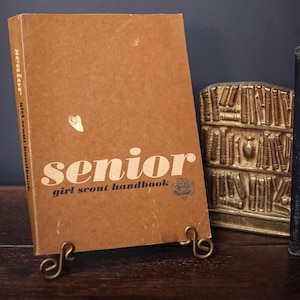 以下が含まれることがあります： 表紙と背表紙に白い文字で「Senior Girl Scout Handbook」と記された茶色のハードカバー本。表紙には小さなグラフィックのエンブレムがあり、金色の金属製スタンドに立てかけられ、その隣には金色のブックエンドが置かれています。