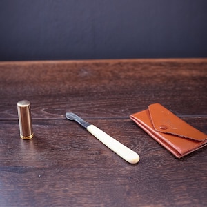 May include: A small, gold-colored metal tube, a white handled, metal cuticle pusher, and a brown leather wallet on a dark wood surface.