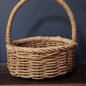 May include: A woven wicker basket with a single handle. The basket is made of natural brown rope and has a round shape.
