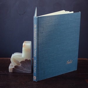 May include: A teal hardcover book titled "The Technique of Landscape Painting" by Frederic Taubes, with a gold embossed signature. The book is propped up by a white and brown marble bookend. The book is on a dark wooden surface.