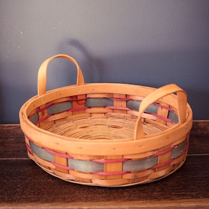 May include: A woven basket with two handles, featuring a light brown base with a blue and pink striped pattern.