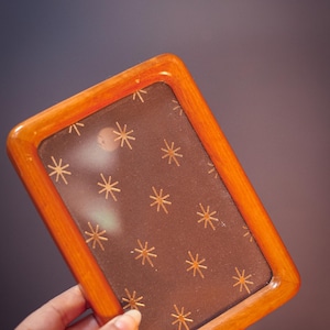 Puede incluir: Un marco de fotos rectangular con un borde de madera de color marrón claro pulido. El panel central es una superficie de color marrón oscuro decorada con un patrón repetitivo de pequeñas estrellas doradas de ocho puntas en cuadrícula.