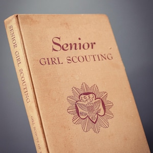 以下が含まれることがあります： Senior GIRL SCOUTINGというタイトルのヴィンテージのベージュ色のハードカバー本。表紙にはマルーン色の文字とエンブレムが描かれています。背表紙にもSenior GIRL SCOUTINGというタイトルとGIRL SCOUTS OF THE U.S.A.という文字がマルーン色で記されています。