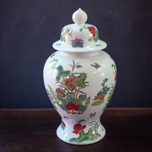 May include: A white ceramic jar with a lid, decorated with a floral pattern in shades of red, orange, green, and purple. The jar has a round body and a wide mouth.