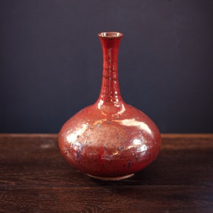 May include: A red ceramic vase with a speckled glaze and a rounded body. The vase has a long, thin neck and a small opening at the top.