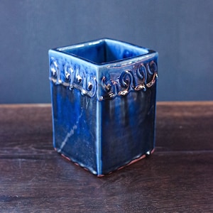 May include: A square blue ceramic vase with a silver-coloured decorative trim around the top edge. The vase is sitting on a dark brown wooden surface.