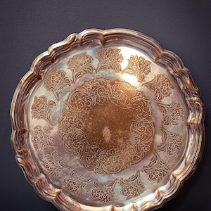 May include: A silver-colored, round tray with a scalloped edge. The tray features an ornate floral pattern across its surface. The design includes detailed flowers and swirling patterns, creating an elegant, vintage aesthetic. The tray is likely made of metal.