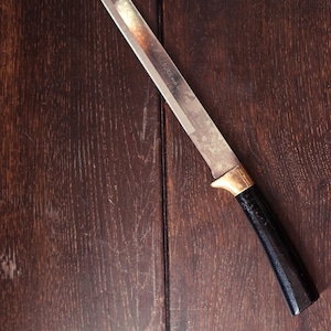 以下が含まれることがあります： 黒いハンドルと金色のアクセントが付いた銀色のチーズナイフ。ナイフは濃い茶色の木製の表面にあります。