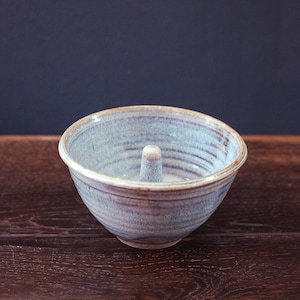 Puede incluir: Un cuenco de cerámica hecho a mano con un esmalte azul claro y un poste cilíndrico central. El cuenco tiene una forma redondeada con un borde ligeramente ensanchado y se asienta sobre una superficie de madera oscura. El diámetro del cuenco es de unos 15 cm.