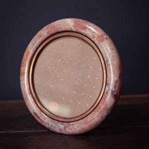 May include: A round photo frame with a pink marble exterior and a copper-colored inner frame. The frame holds a light brown backing with small gold dots. The frame is a decorative item.
