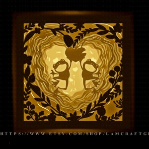 Può includere: Un'opera d'arte in carta a strati con due sagome di rane che tengono un oggetto a forma di cuore. L'opera d'arte è illuminata dall'interno, creando un bagliore caldo e dorato. Il design include ritagli floreali e di foglie.