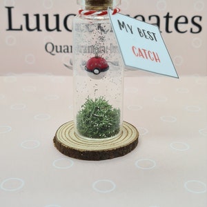 May include: A clear glass bottle with a cork stopper, containing a red and white Pokeball, green moss, and glitter. The bottle sits on a wooden slice and has a tag that reads "MY BEST CATCH".