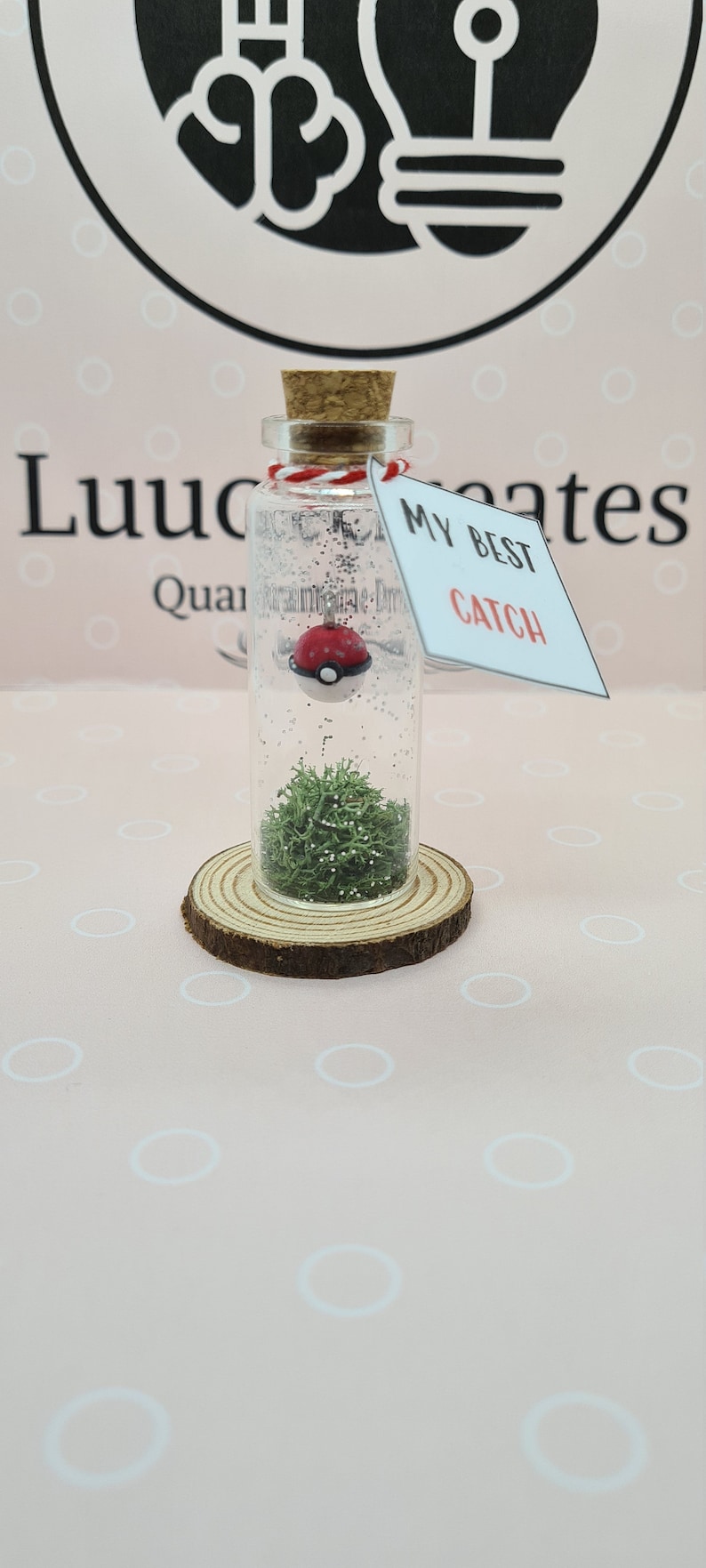 May include: A clear glass bottle with a cork stopper, containing a red and white Pokeball, green moss, and glitter. The bottle sits on a wooden slice and has a tag that reads "MY BEST CATCH".