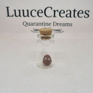 Può includere: Una piccola bottiglia di vetro con tappo di sughero, contenente una figura di argilla marrone con un volto semplice. La bottiglia è posta su uno sfondo chiaro con il testo "LuuceCreates Quarantine Dreams".