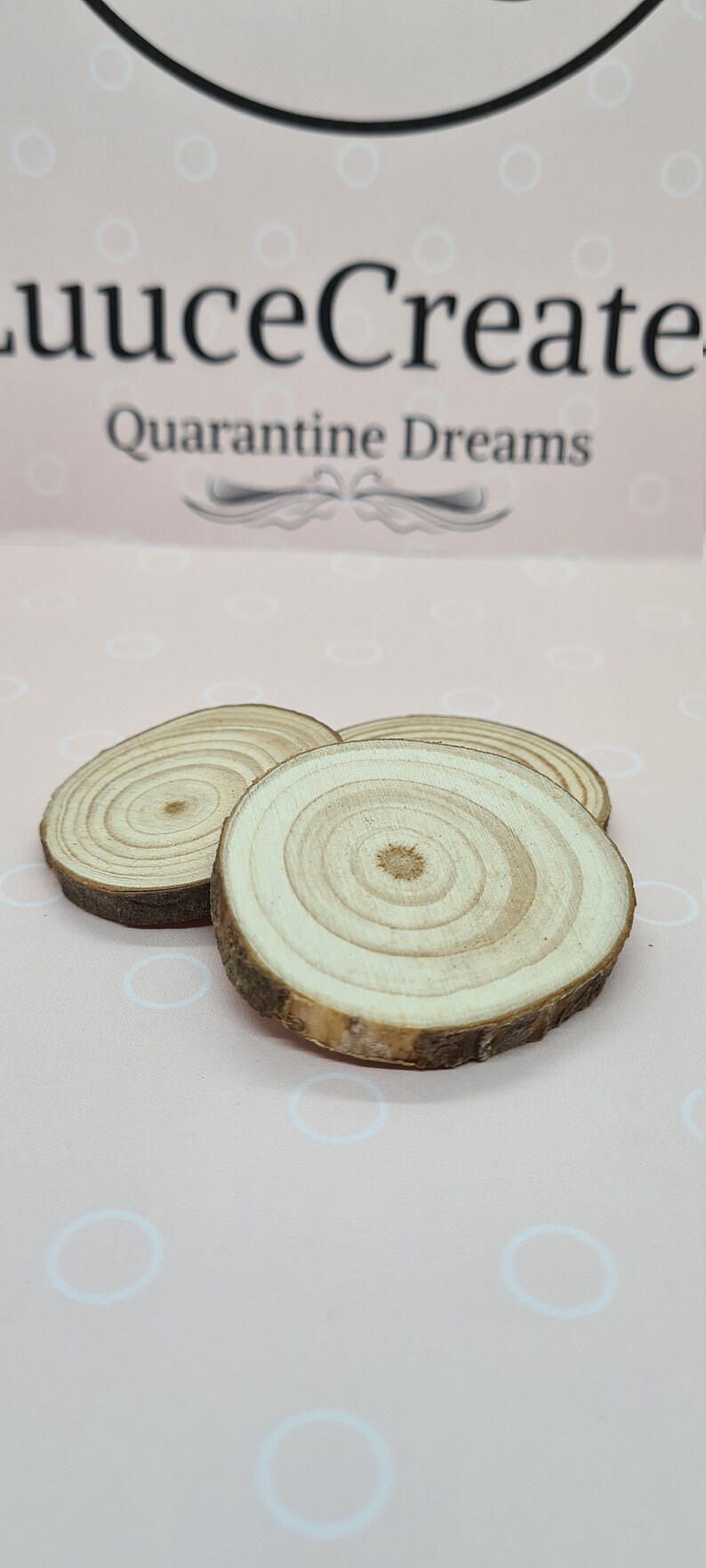 May include: Three small, round wooden slices with visible growth rings and bark edges. The slices are light brown and tan, arranged on a pink background with the text "luuceCreate Quarantine Dreams" at the top.