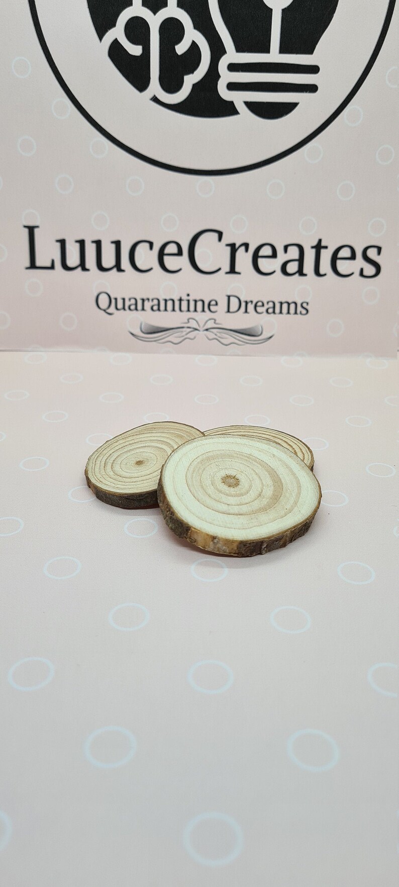 May include: Three small, round wooden slices with visible growth rings and bark edges. The slices are arranged on a pink surface with a pattern of white circles. The text "LuuuceCreates Quarantine Dreams" is visible in the background.