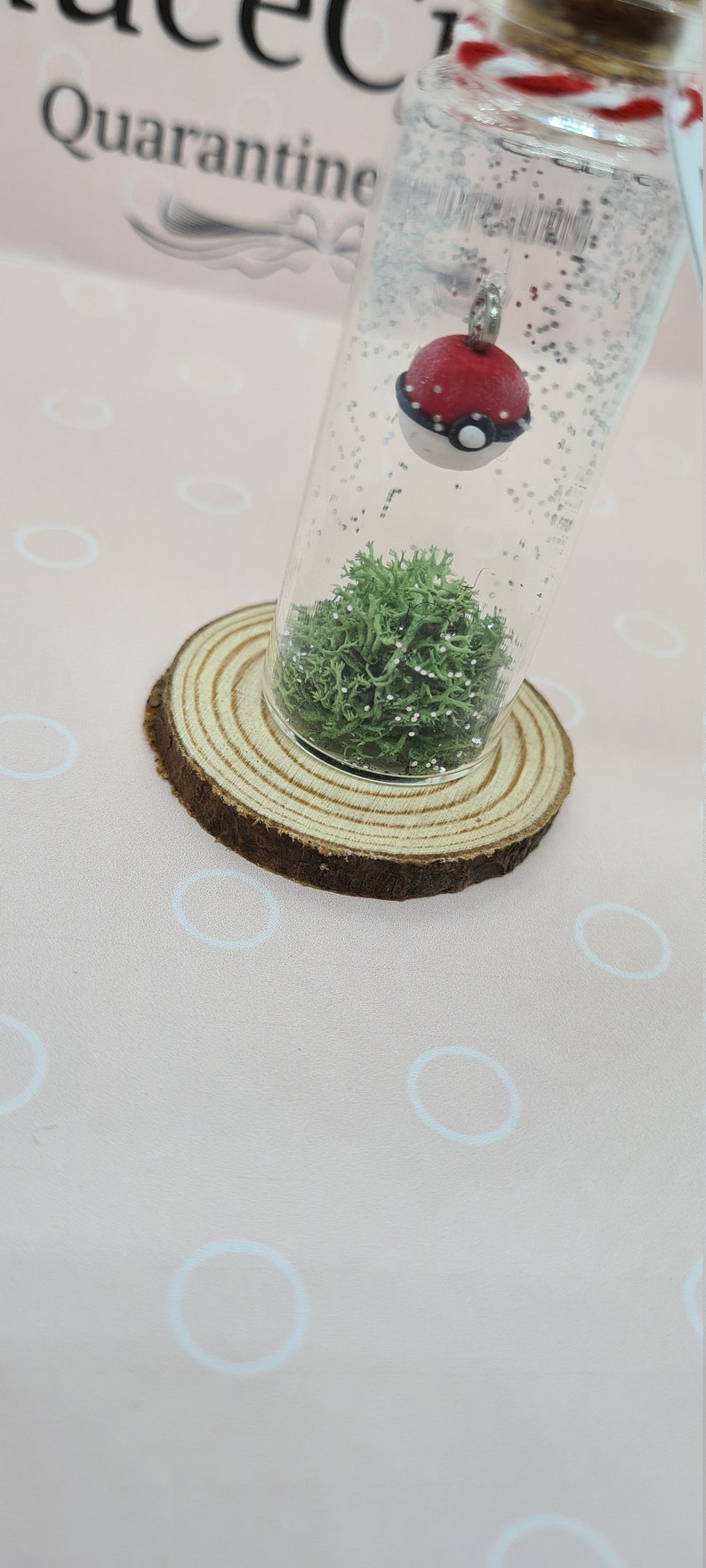 May include: A clear glass vial with a red and white charm resembling a Pok&eacute; Ball, suspended in a glitter-filled liquid. The vial rests on a wooden slice, with green moss at the bottom. The background is a light pink surface.