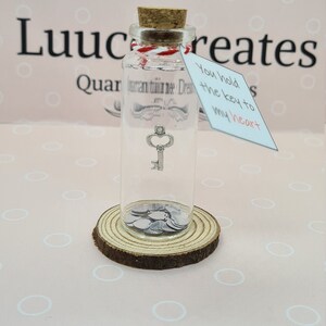 May include: A clear glass bottle with a cork stopper, containing a small silver key charm and silver heart-shaped confetti. A tag reads "You hold the key to my heart". The bottle sits on a wooden slice.