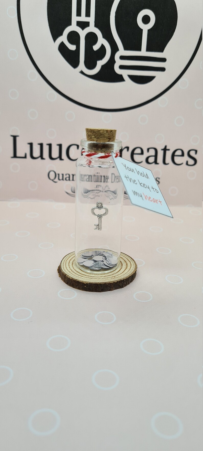 May include: A clear glass bottle with a cork stopper, containing a small silver key charm and silver heart-shaped confetti. A tag reads "You hold the key to my heart". The bottle sits on a wooden slice.