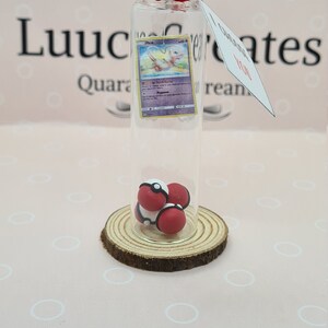 May include: A clear glass bottle with a cork stopper, containing a Pokemon card and three red and white balls. The bottle sits on a wooden slice. A tag with the word "Love" hangs from the bottle.