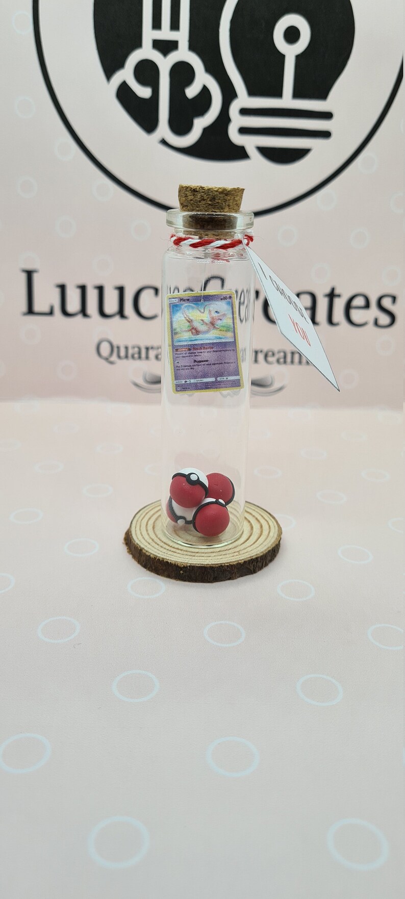 May include: A clear glass bottle with a cork stopper, containing a Pokemon card and three red and white balls. The bottle sits on a wooden slice. A tag with the word "Love" hangs from the bottle.