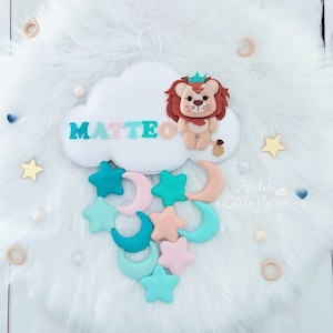 Può includere: Una nuvola di feltro bianco con il nome "MATTEO" in lettere colorate e un leone di feltro con una corona. La nuvola è appesa a una corda e ha una ghirlanda di stelle e lune di feltro appesa sotto.