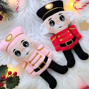 Peut inclure: Deux poupées casse-noisettes en peluche, une rose et une rouge, sont posées sur une surface blanche et duveteuse. La poupée rose a des détails dorés, tandis que la poupée rouge a des bottes noires. Des décorations de Noël, dont des cannes en sucre et une boule rouge, entourent les poupées.