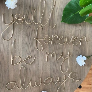 May include: Gold wire letters spelling out the phrase "you'll forever be my always..." on a wooden surface. White flowers are scattered around the letters.