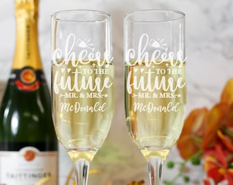 May include: Two champagne flutes with the text "Cheers to the future Mr. & Mrs. McDonald" etched on them. The flutes are filled with champagne and are sitting on a white surface.