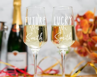 May include: Two champagne flutes filled with a bubbly beverage. One glass reads "FUTURE Mrs Smith", the other reads "LUCKY Mr Smith". A bottle of champagne and a cocktail shaker are in the background. The flutes are on a white surface.