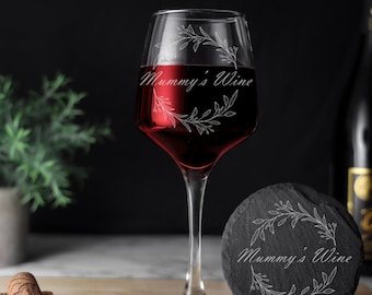 May include: A clear wine glass with a stem and a wide bowl. The glass is etched with a floral design and the words "Mummy's Wine". The glass is sitting on a black slate coaster with the same design and text.