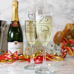 May include: Two champagne flutes with engraved hearts and the text "Richard 25th Anniversary" and "25th Anniversary Christi" on them. The flutes are filled with champagne and are sitting on a white surface with red and gold ribbon.