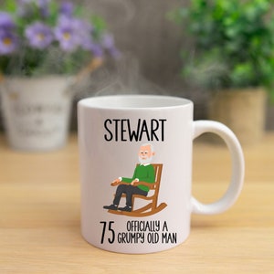 May include: White ceramic mug with the name "STEWART" in black text. The mug features an illustration of an elderly man in a rocking chair, with the text "75 OFFICIALLY A GRUMPY OLD MAN".