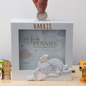 May include: White wooden money box with a clear plastic frame. The frame has a blue background with white clouds and stars. The text "my first pennies" is printed in gold glitter. A cartoon image of a sleeping elephant is in the frame. The money box has a slot for coins and the name "HARRIS" is printed on the top.