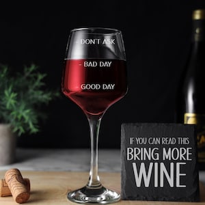 May include: A clear wine glass with the words "Don't Ask", "Bad Day", and "Good Day" etched on the side. The glass is filled with red wine. A black slate coaster with the text "If you can read this bring more wine" is in the foreground.