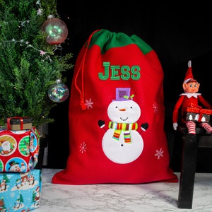 May include: Red and green Christmas gift bag with a snowman design and the name "Jess" embroidered on the front.