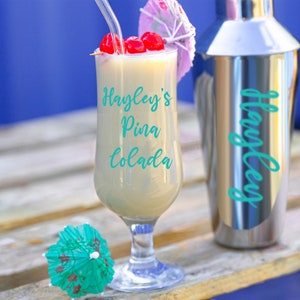 Puede incluir: Un vaso de Pina Colada de Hayley con una sombrilla de papel rosa y blanca y cerezas maraschino. La bebida est&aacute; en un vaso alto con tallo y base transparente. El vaso est&aacute; sobre una superficie de madera.