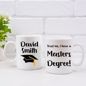 Peut inclure: Deux tasses en céramique blanche avec du texte noir et un motif de chapeau de graduation. La première tasse dit "David Smith" et la deuxième tasse dit "Croyez-moi, j'ai une maîtrise !"
