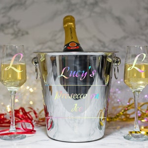 May include: A silver champagne bucket with the text "Lucy's Prosecco on Ice" in rainbow iridescent lettering. Two champagne flutes with the letter "L" in rainbow iridescent lettering are on either side of the bucket.