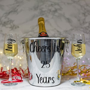 May include: A silver champagne bucket with the text "Cheers to 25 Years" on it. Two champagne flutes are on either side of the bucket, one with the name "Amy" and the other with the name "John".