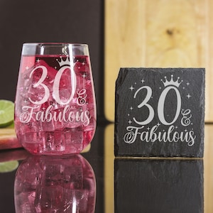 May include: A black slate coaster with the text "30 & Fabulous" in white with a crown above the number 30. A clear glass with a pink drink inside has the same text etched on it.