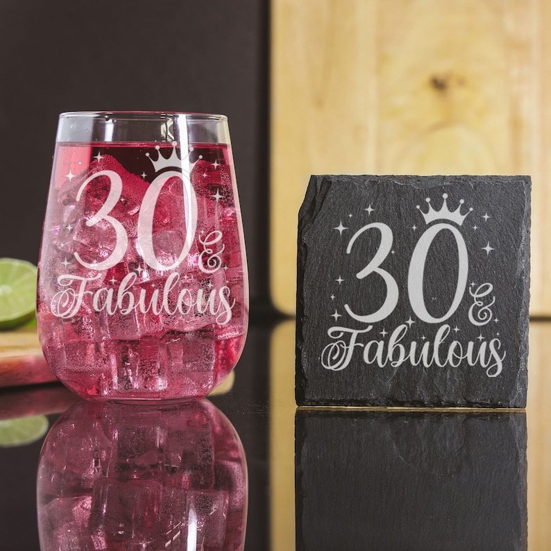 May include: A black slate coaster with the text "30 & Fabulous" in white with a crown above the number 30. A clear glass with a pink drink inside has the same text etched on it.