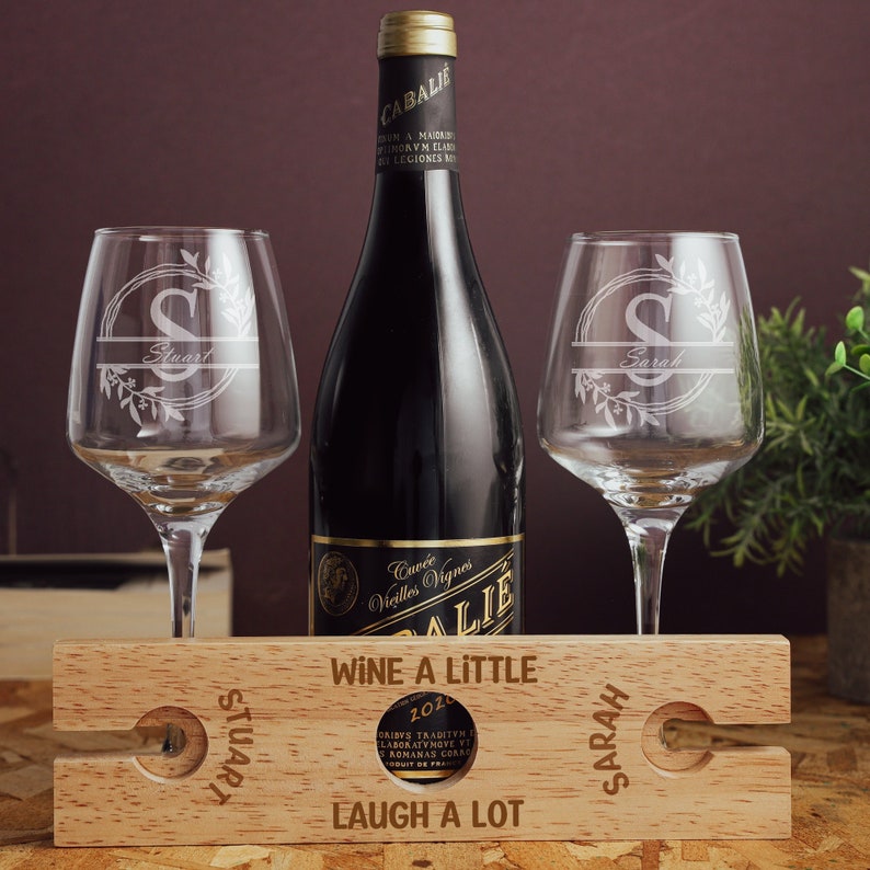 Puede incluir: Un soporte de madera para botellas de vino con dos copas de vino. Las copas est&aacute;n grabadas con las letras "S" y los nombres "Stuart" y "Sarah". El soporte tiene el texto "Wine a little, Laugh a lot" y los nombres "Stuart" y "Sarah" grabados.