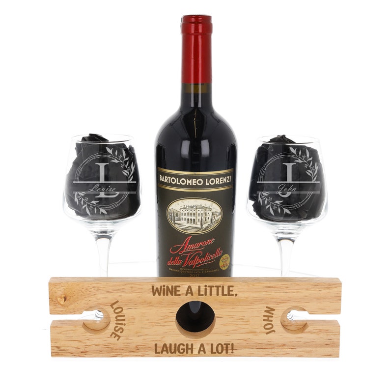 Puede incluir: Un estante de vino de madera con dos vasos y una botella de vino. Los vasos est&aacute;n grabados con los nombres "Louise" y "John". El estante tiene el texto "Wine a little, laugh a lot!" grabado.