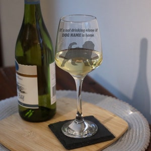 May include: A clear wine glass with a silhouette of a dog and the text "It's not drinking alone if DOG NAME is home." The glass is filled with white wine and is sitting on a black coaster on a wooden surface.