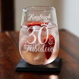 May include: A clear glass wine glass with the text "Kayleigh 30 & Fabulous" etched on the side. The glass is filled with a pink drink and ice.
