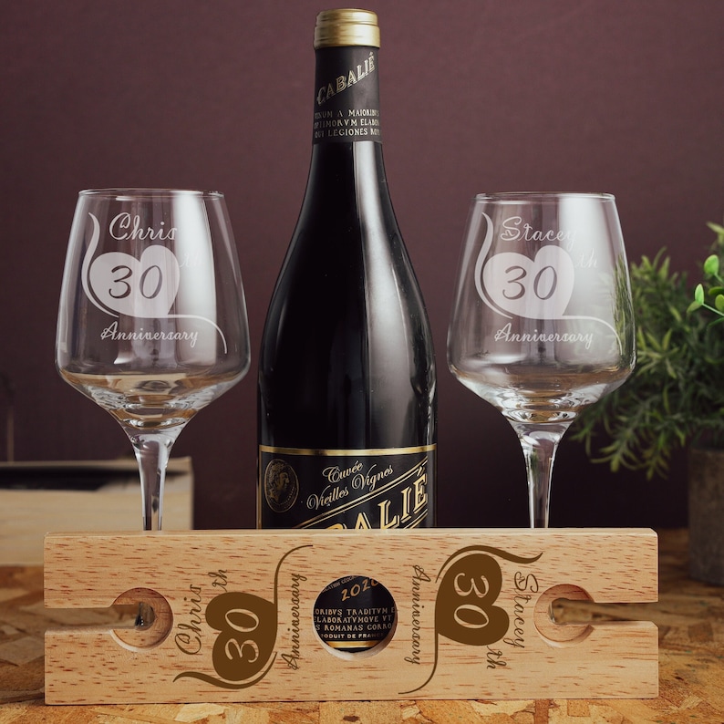 May include: A wooden wine bottle holder with two wine glasses. The glasses are engraved with "Chris 30th Anniversary" and "Stacey 30th Anniversary". The bottle holder is engraved with "Chris 30th Anniversary" and "Stacey 30th Anniversary".