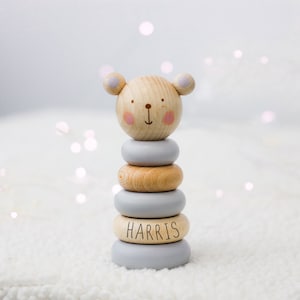 May include: A wooden stacking toy with a bear face on top. The toy is made of natural wood and painted in light blue and brown. The bottom ring has the name "HARRIS" written on it.